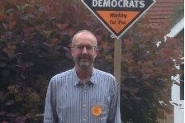 Image of the candidate Graham Burr, wearing a striped search, with an orange badge, standing next to a Liberal Democrat sign