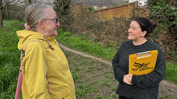 A person with a yellow clipboard, showing the liberal democrat logo, smiling and having a conversation with a second person on a grassy footpath .