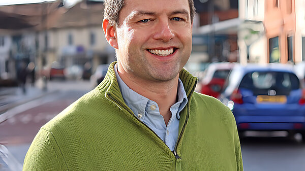 A picture of Chris Coghlan MP, in the sunshine on Horley high street, wearing a blue shirt and white zip up top, smiling towards the camera