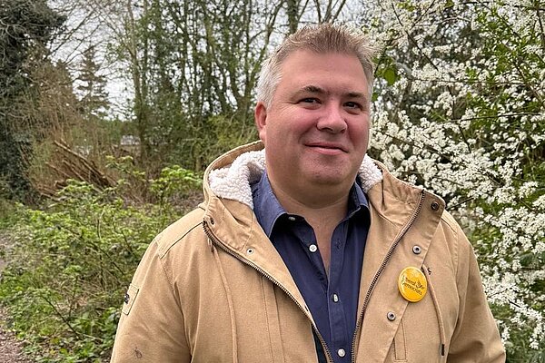 A picture of a man with a tan jacket, blue shirt and lib dem lapel