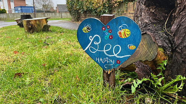 A picture of the edge of a park. Paths can be seen in the background. A blue heart shaped sign saying "Bee happy" can be seen in the foreground, with a tree behind it. The picture is taken from low donw.